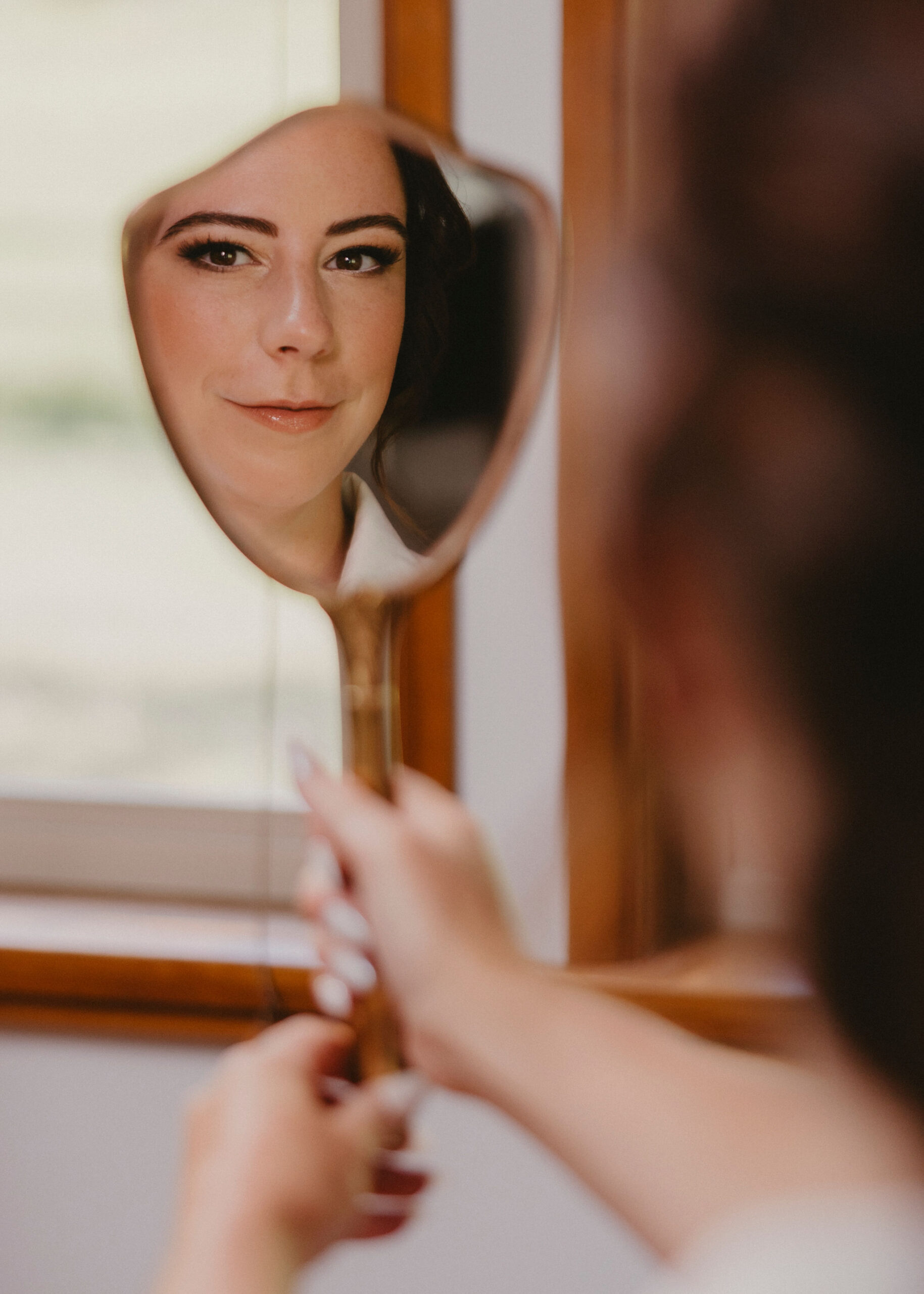 Bride looking at her makeup in a vintage mirror. Photographer capturing the reflection in the mirror.